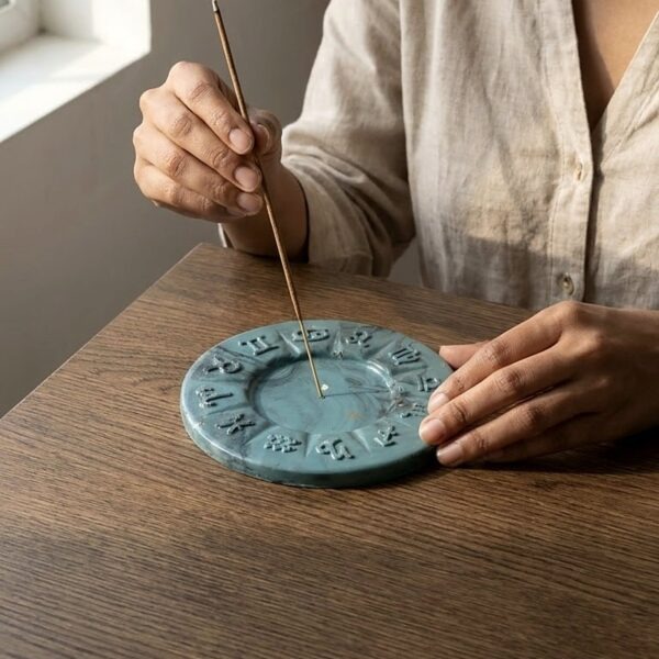 Teal marbled jesmonite incense holder with raised zodiac signs, with an agarbatti being placed in the centre on a wooden table